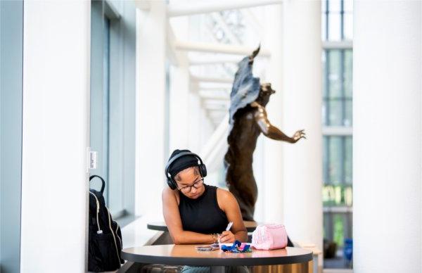  A college student wearing headphones writes in a journal while in a library. 